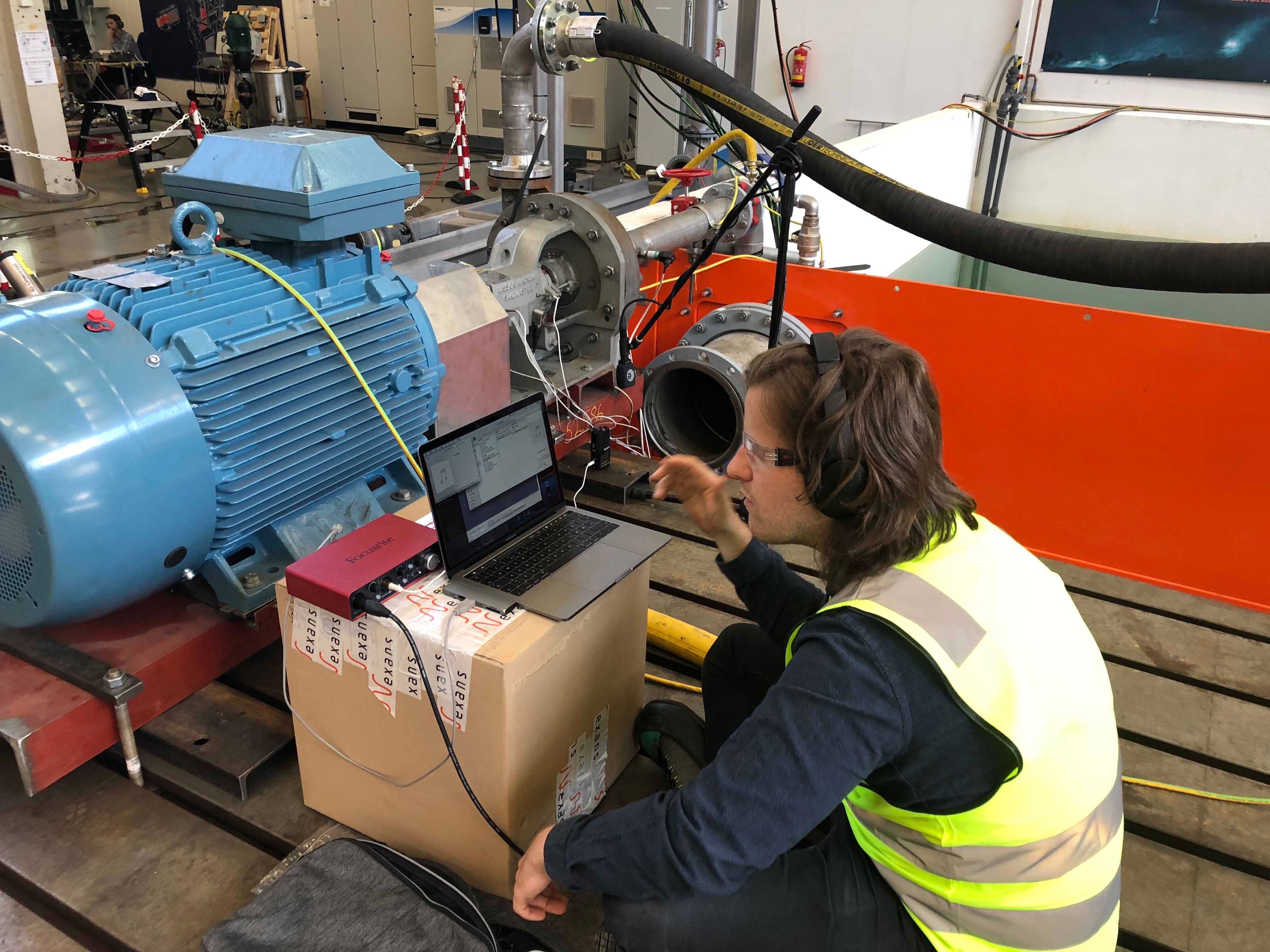 Man sitting in front of his microphone equipped laptop, in front of a large pump, doing experimental analysis of audio data