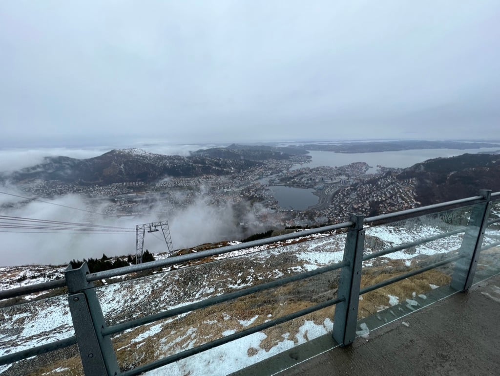 Utsikt fra gondolbaneplattformen på Ulriken, med en bitte liten Bergen by i bakgrunnen; i forgrunnen snaufjell med snø og et tårn fra kabelbanen